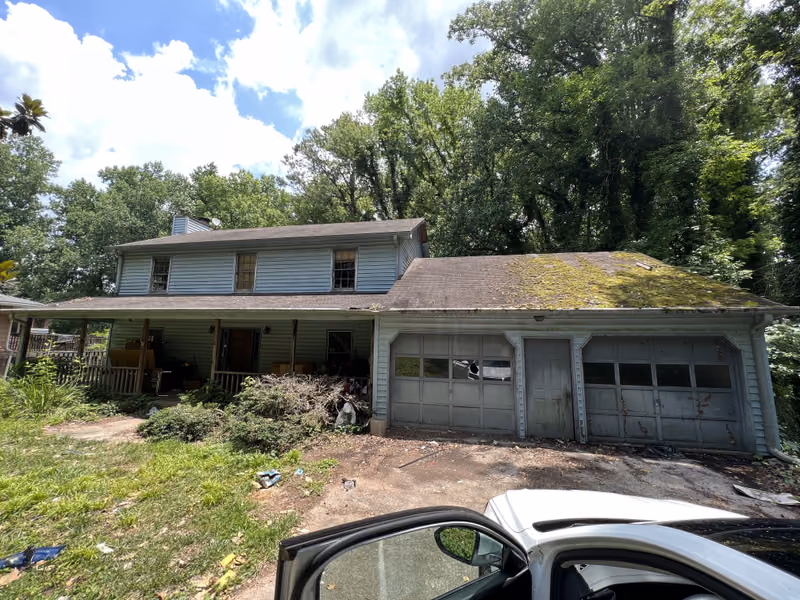 Two-story house with moss-covered garage roof and overgrown landscaping, bought in any condition